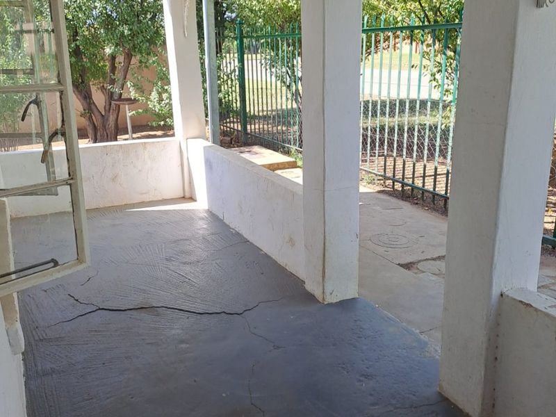 A covered outdoor patio area featuring concrete flooring with visible cracks, supported by structural pillars and enclosed by a metal security fence.