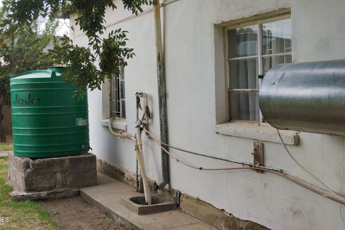 Rear exterior view showing a water storage tank on a concrete plinth, external plumbing, and a wall-mounted geyser unit.