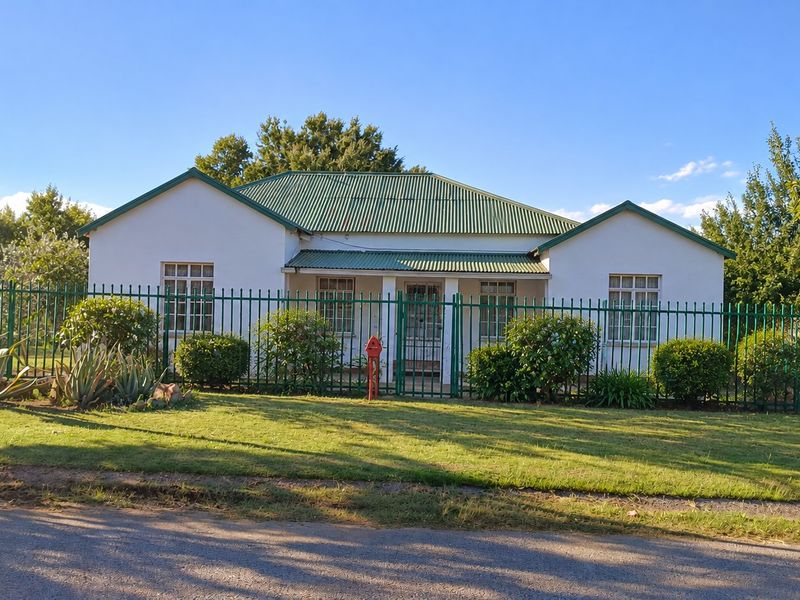 Front elevation of a single-story house featuring a green corrugated metal roof, covered porch, and secure perimeter fencing.