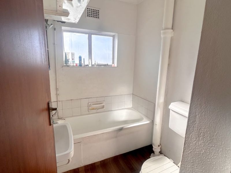 A compact, dated bathroom featuring a bathtub, toilet, and wall-mounted basin, requiring renovation to modernize the finishes and plumbing presentation.