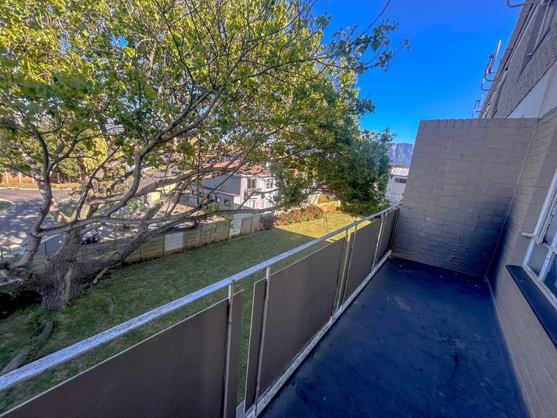 A private balcony featuring a metal balustrade and brick wall, overlooking a shared garden area with mature trees.