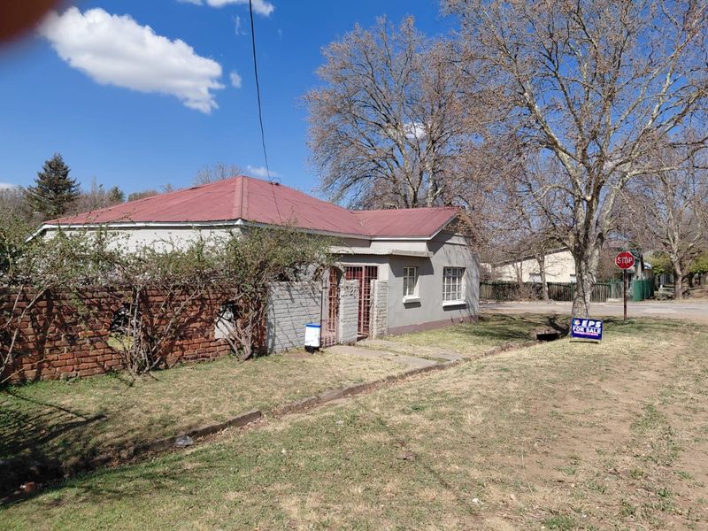 Front view of a single-story house featuring a corrugated metal roof, brick boundary wall, and metal security gate, set on a corner plot.