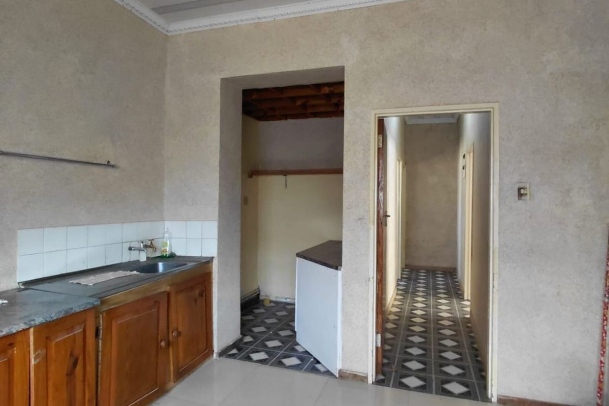 A dated kitchen area featuring wooden cabinetry, tiled flooring, and a panelled ceiling, leading into a hallway and adjacent utility space.