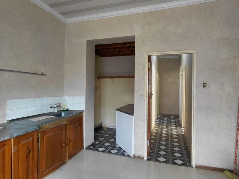 A dated kitchen area featuring wooden cabinetry, tiled flooring, and a panelled ceiling, leading into a hallway and adjacent utility space.