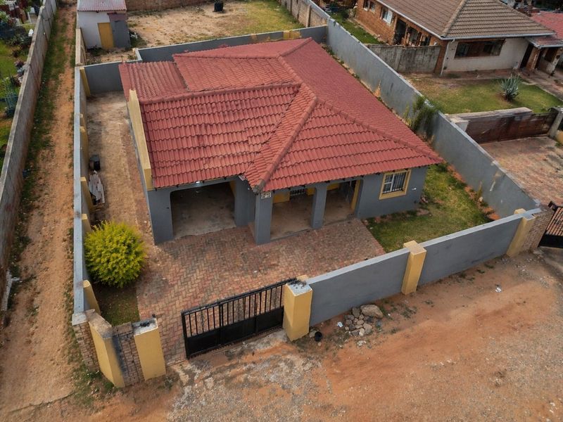 Aerial view of a single-story house featuring a tiled roof, paved driveway, and enclosed perimeter wall with a metal gate.