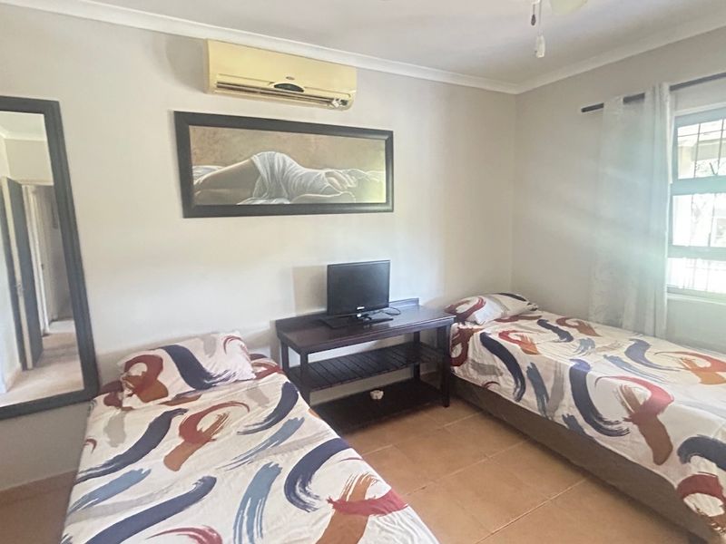 A functional bedroom featuring tiled flooring, a wall-mounted air conditioning unit, and a window fitted with security bars.