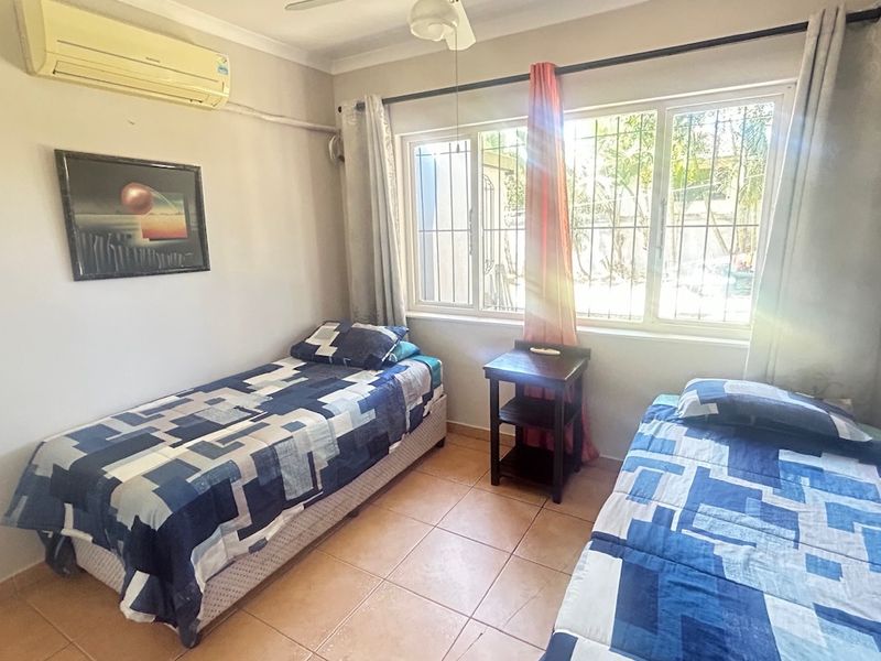 A functional bedroom featuring tiled flooring, a wall-mounted split-system air conditioner, a ceiling fan, and large windows fitted with security bars.