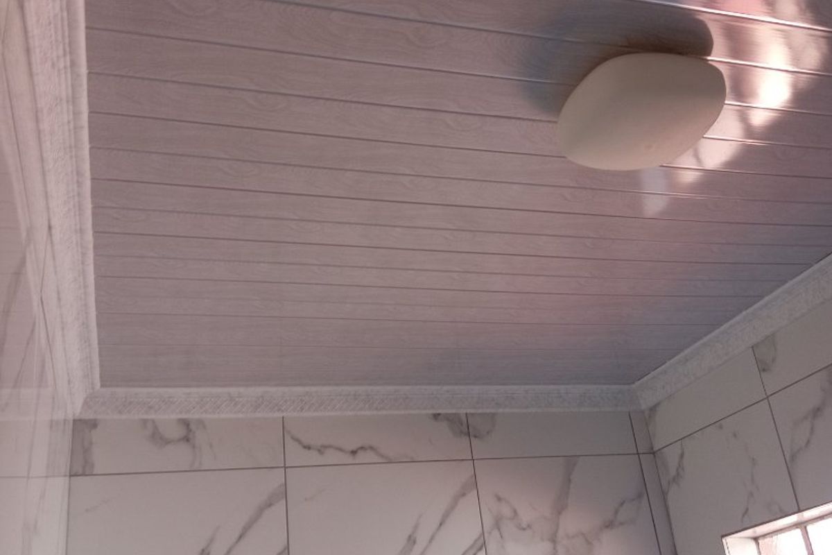 A clean bathroom interior featuring modern marble-look wall tiles, a wood-effect PVC ceiling panel finish, and decorative cornice detailing.