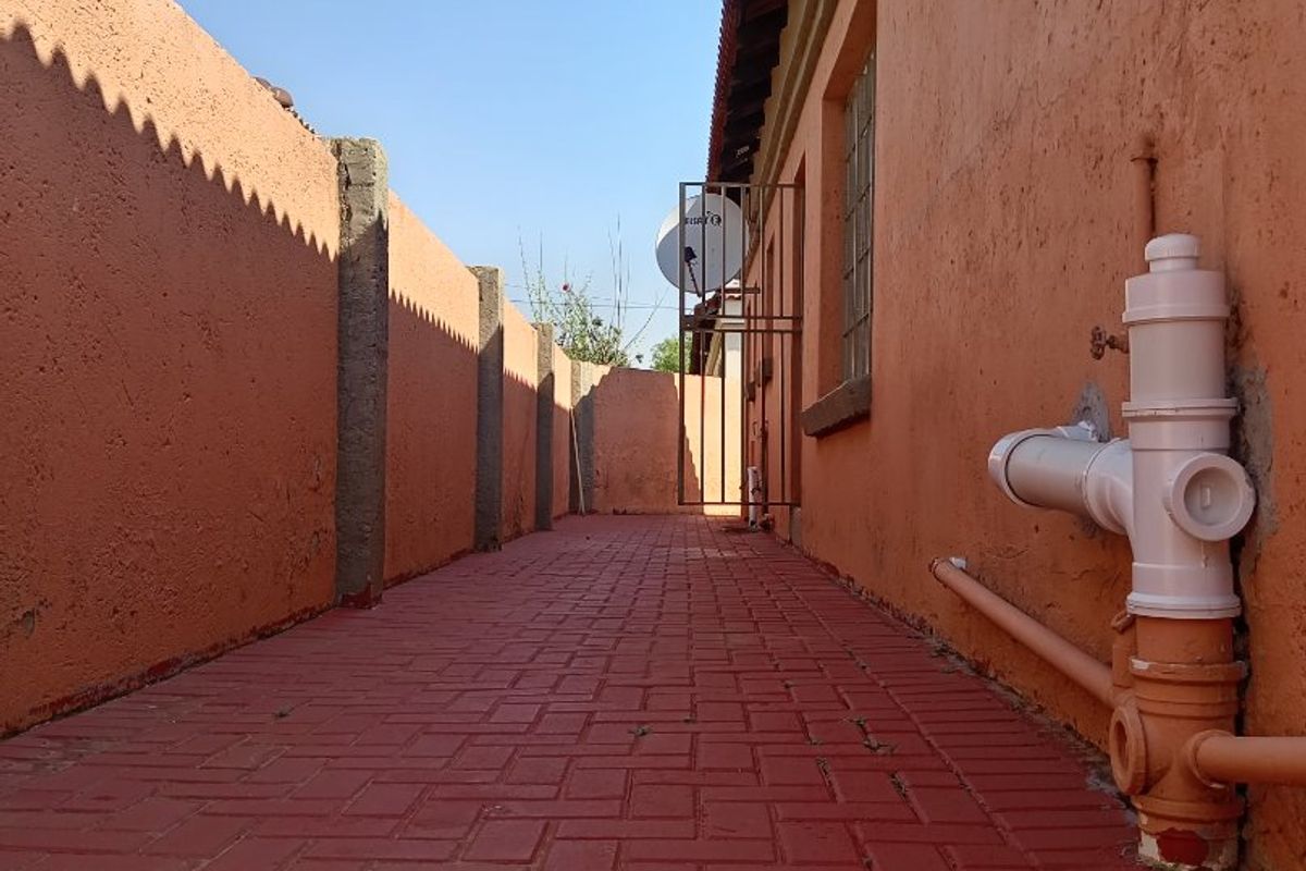 A side passage or rear exterior area featuring red brick paving, boundary walls, and exposed external plumbing infrastructure.
