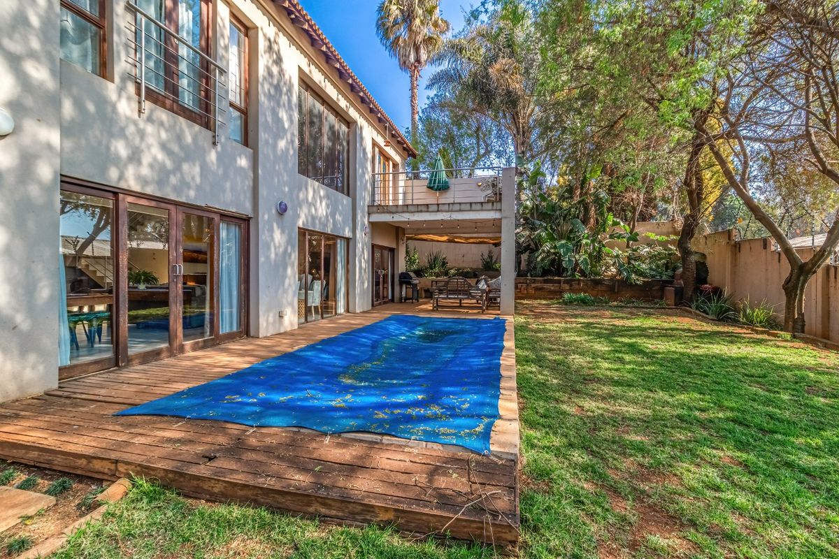 Rear exterior view featuring a wooden deck, swimming pool with cover, balcony, and a spacious garden area with mature trees.