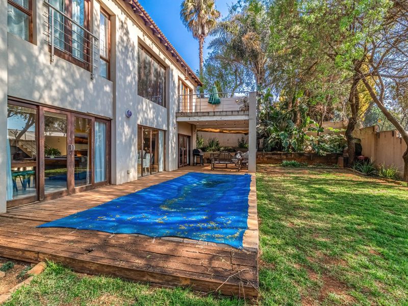 Rear exterior view featuring a wooden deck, swimming pool with cover, balcony, and a spacious garden area with mature trees.