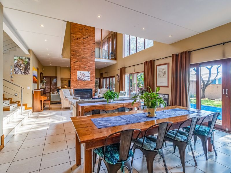 Spacious open-plan living and dining area featuring a double-height brick fireplace, tiled flooring, and large windows providing natural light and garden access.
