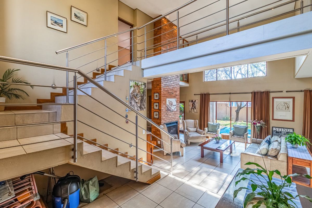 Spacious double-volume living area featuring a prominent brick fireplace, tiled flooring, and a modern staircase with stainless steel balustrades leading to a mezzanine level.