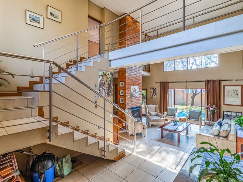 Spacious double-volume living area featuring a prominent brick fireplace, tiled flooring, and a modern staircase with stainless steel balustrades leading to a mezzanine level.
