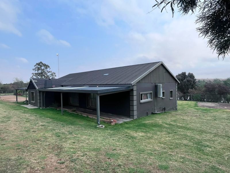 Rear view of a farm-style dwelling featuring a corrugated metal roof, covered patio area, and external geyser unit set on a large grassed plot.