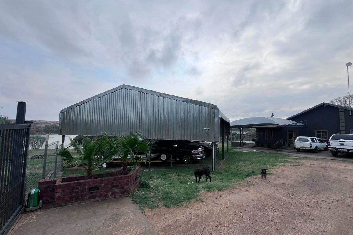 Rear exterior view of the property featuring a large corrugated metal carport, shade sail structure, and brick landscaping elements.