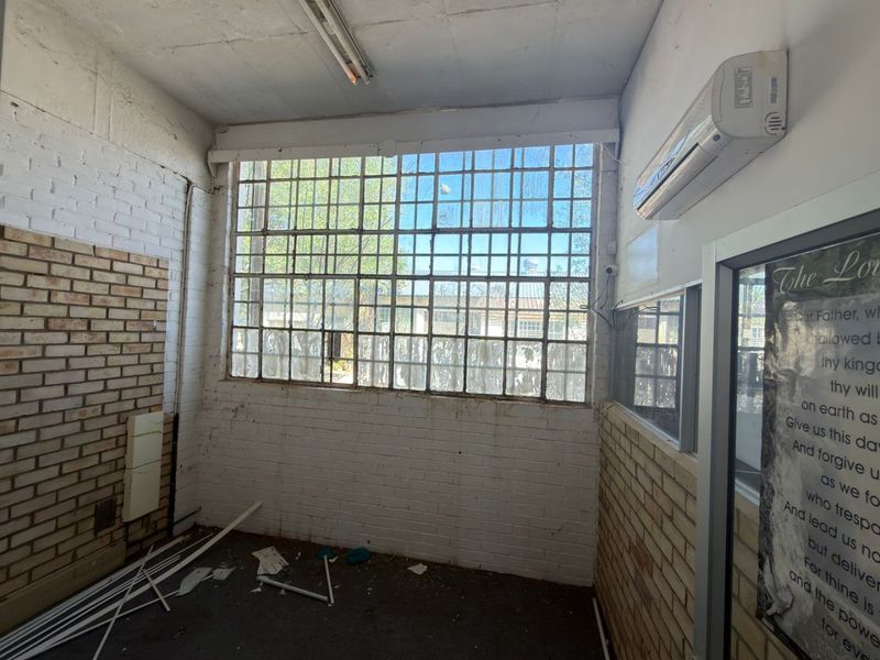An empty office space featuring industrial steel-framed windows, exposed brick walls, and a wall-mounted air conditioning unit, requiring refurbishment.