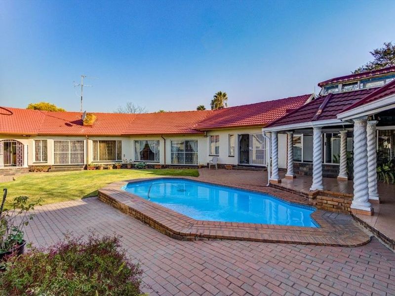 Spacious rear exterior featuring a swimming pool, brick paved patio area, and covered veranda with decorative columns, set against the main house structure.