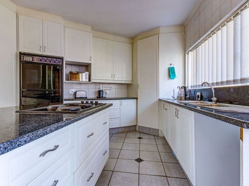 A functional kitchen featuring white cabinetry, granite countertops, a built-in oven, and an electric hob, with tiled flooring and natural light from vertical blinds.