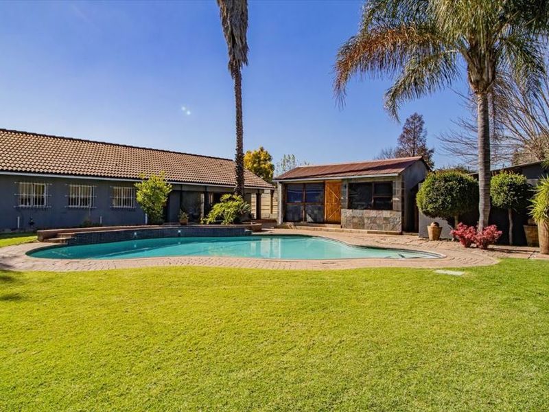 Spacious backyard featuring a large swimming pool with paved surround, manicured lawn, and a detached outbuilding under a tiled roof.