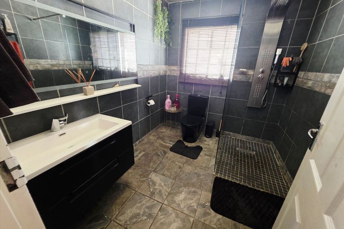 Modern bathroom featuring dark tiled walls and flooring, a wall-mounted vanity with integrated basin, and a walk-in shower with a stainless steel panel.