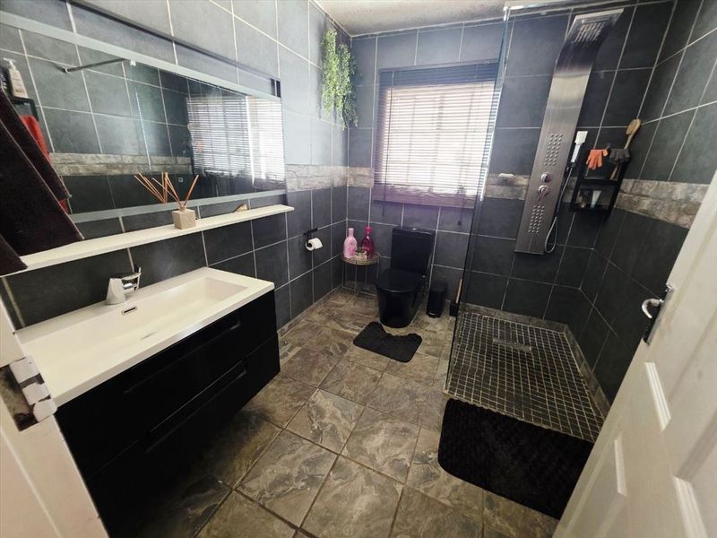 Modern bathroom featuring dark tiled walls and flooring, a wall-mounted vanity with integrated basin, and a walk-in shower with a stainless steel panel.