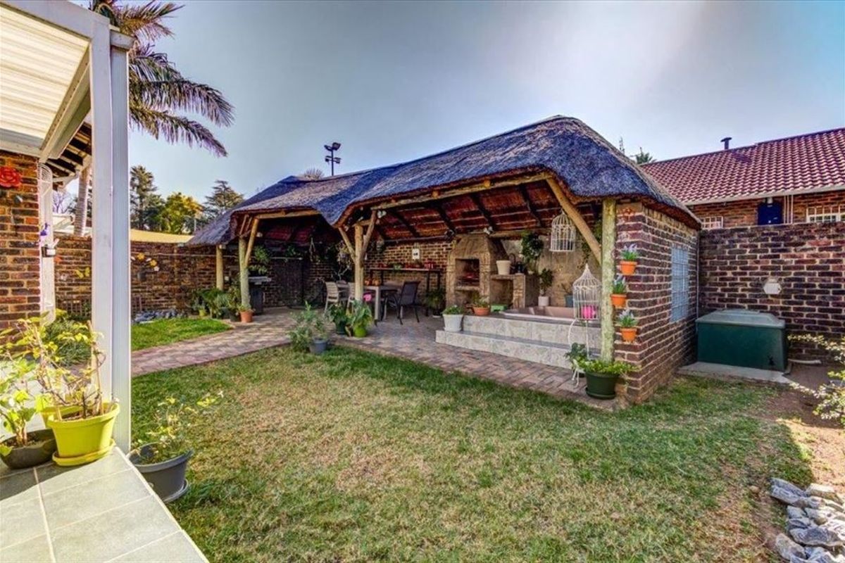 A rear garden featuring a thatched lapa with a built-in braai, brick paving, and a raised platform area, perfect for outdoor entertaining.