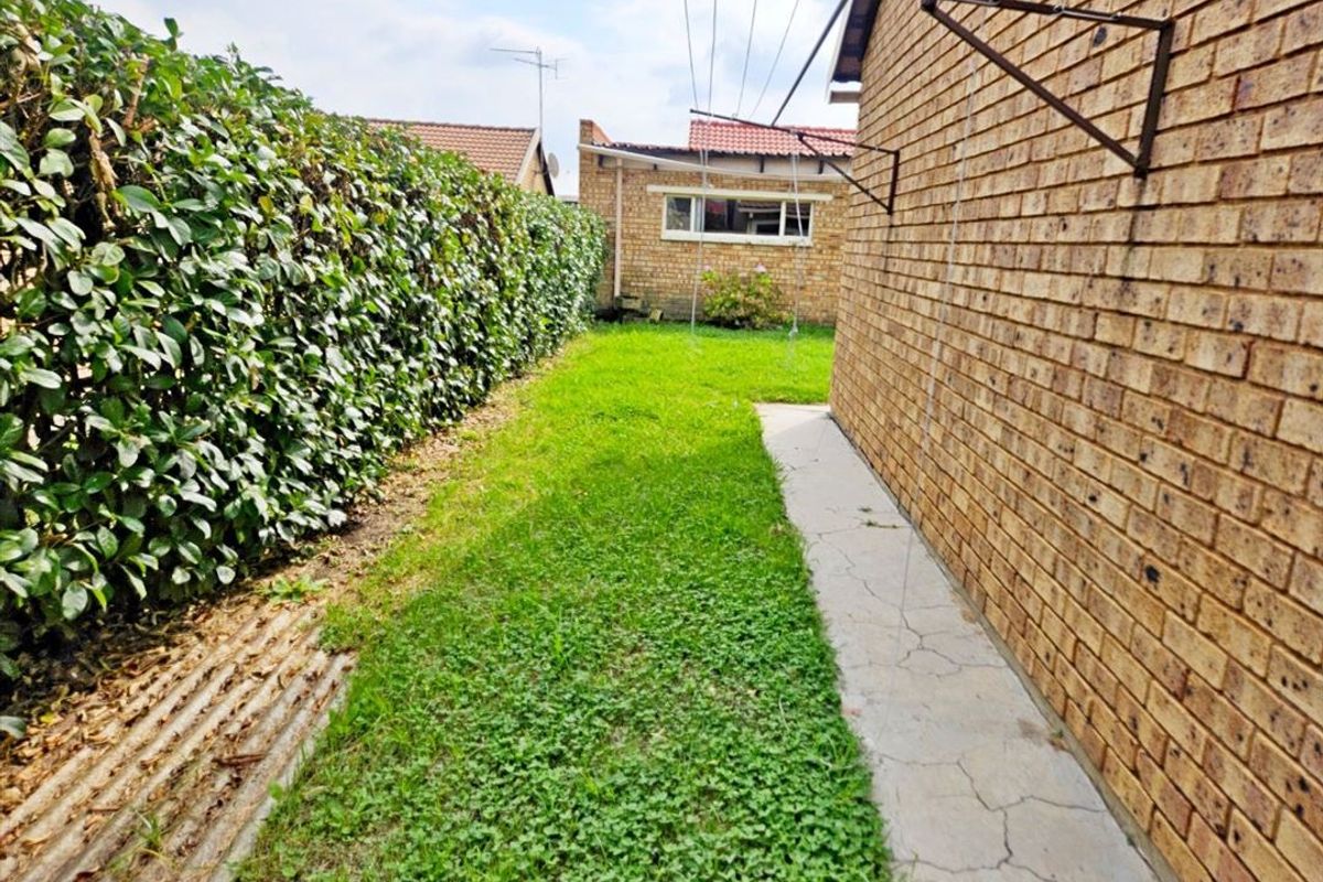 A rear exterior view of the townhouse featuring a brick wall, a small lawn area, a hedge boundary, and a wall-mounted washing line.