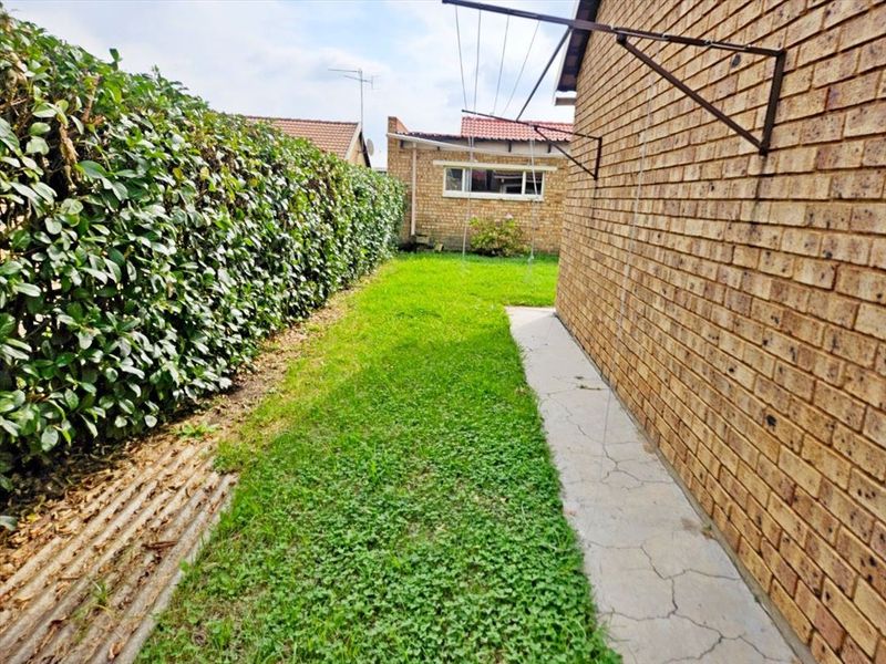A rear exterior view of the townhouse featuring a brick wall, a small lawn area, a hedge boundary, and a wall-mounted washing line.
