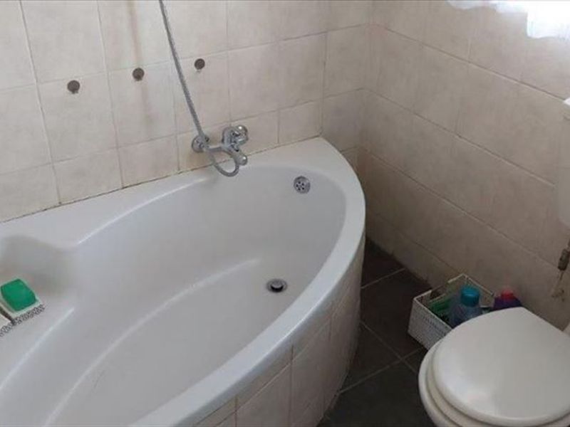 A compact bathroom featuring a corner bathtub, wall-mounted shower mixer, and a standard close-coupled toilet with tiled walls and flooring.