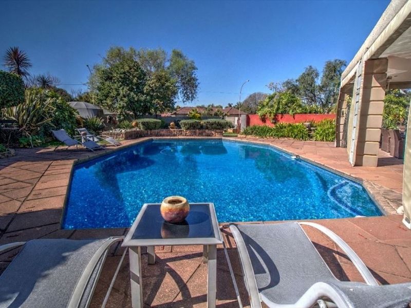 A well-maintained outdoor swimming pool area featuring a paved deck, stone retaining wall, and an adjacent covered patio space.