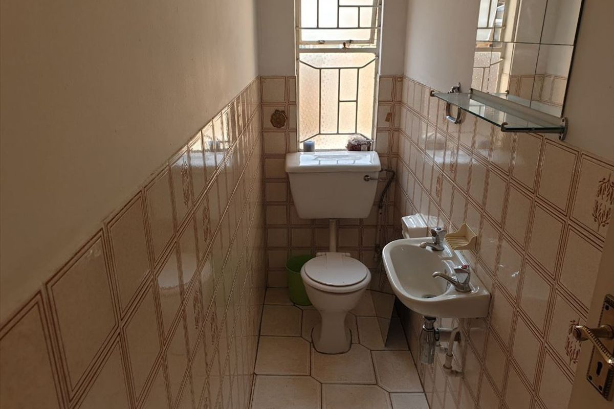 A compact guest bathroom featuring a wall-mounted basin, toilet, and dated ceramic wall and floor tiling with security bars on the window.