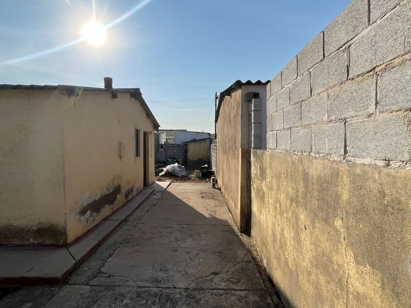 A narrow exterior side passage featuring a concrete walkway between two structures, enclosed by a cinder block boundary wall with visible signs of weathering.