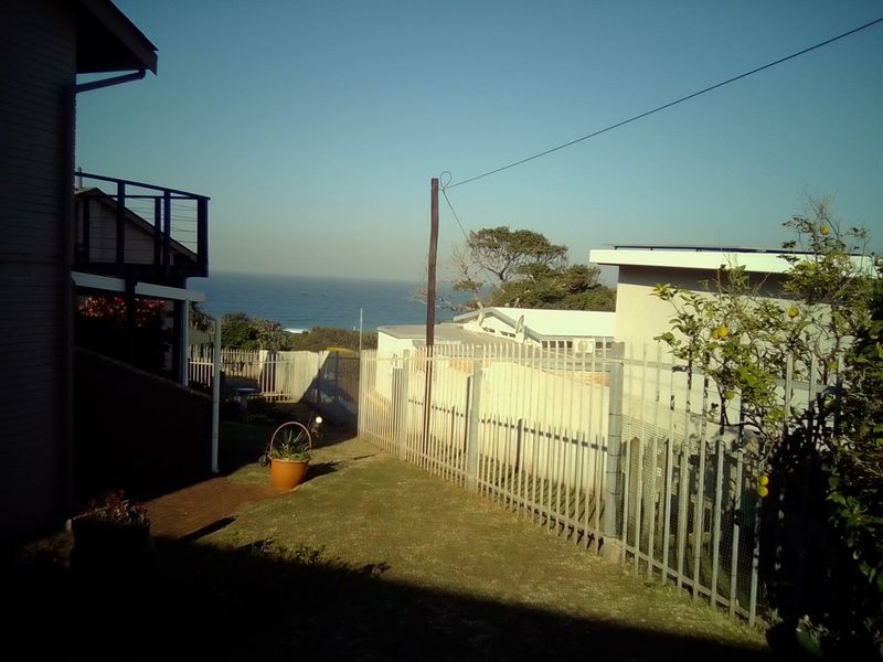 A rear exterior view of the property featuring a lawn, metal security fencing, and a paved walkway with a distant ocean view.