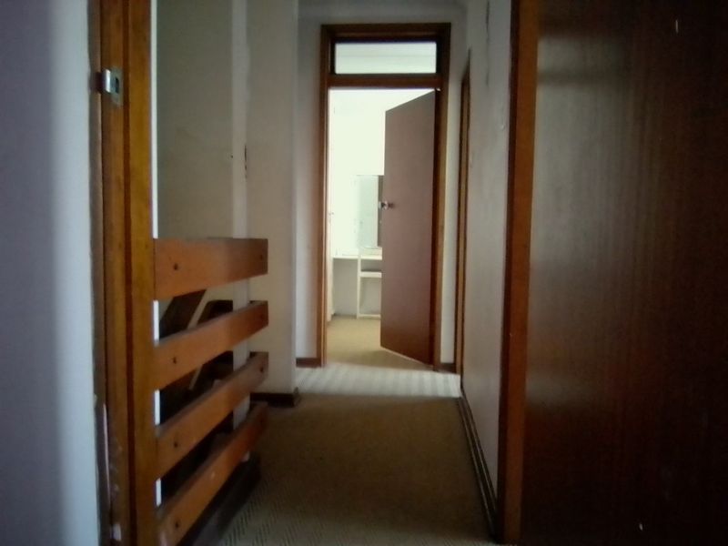 A narrow interior hallway featuring wooden door frames, a wooden balustrade, and carpeted flooring, leading towards an adjacent room.