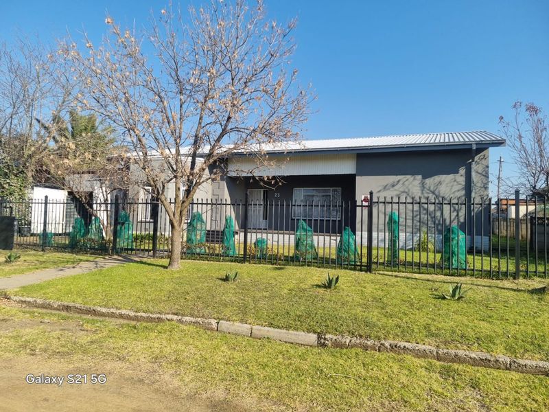 Front exterior view of a single-story house featuring a grey facade, corrugated metal roof, and secure metal perimeter fencing.
