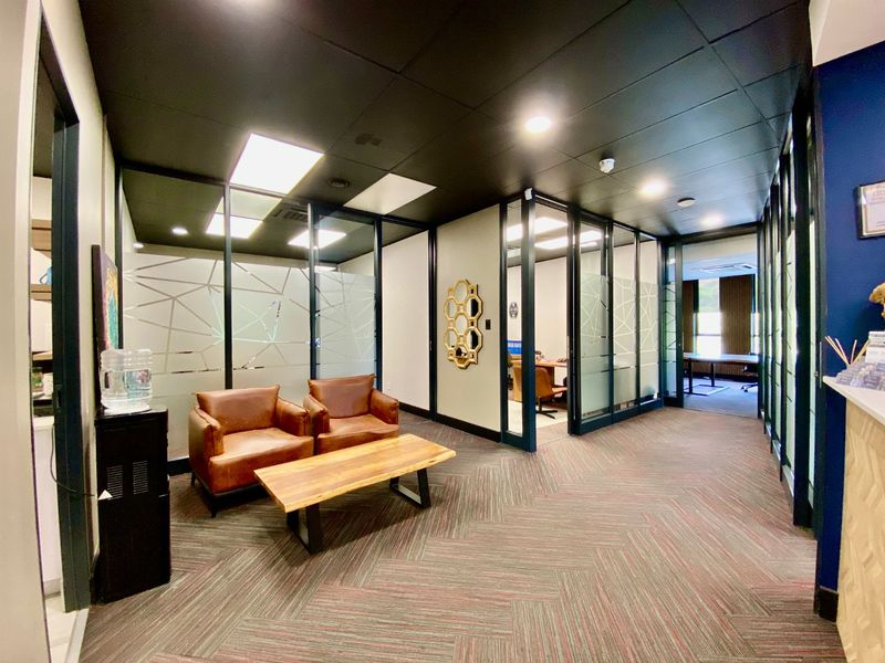 A modern commercial office hallway featuring glass-partitioned private offices, recessed ceiling lighting, and patterned commercial carpeting.
