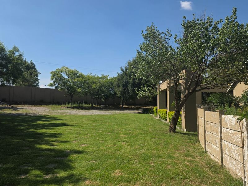 A spacious backyard featuring a large lawn, mature trees, and a covered patio area, enclosed by a precast concrete boundary wall.