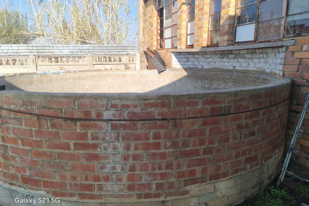 A rear exterior view featuring a circular brick water tank structure adjacent to a brick building with aged steel-framed windows and perimeter security fencing.