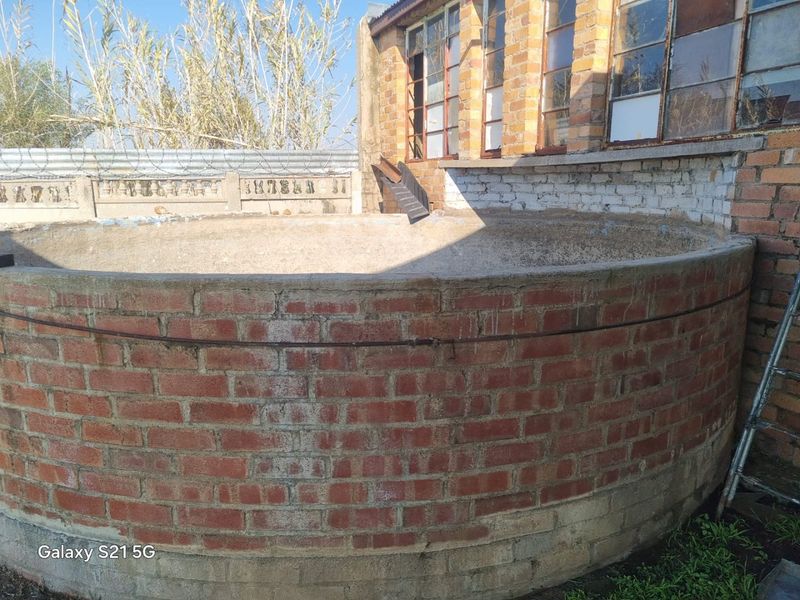 A rear exterior view featuring a circular brick water tank structure adjacent to a brick building with aged steel-framed windows and perimeter security fencing.