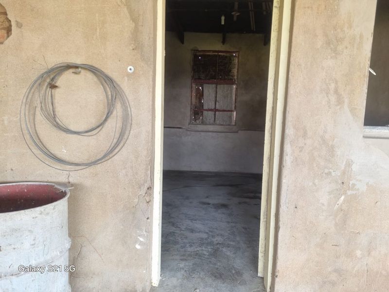 A vacant utility space featuring concrete flooring, exposed ceiling beams, and a weathered metal window frame, requiring significant renovation and repair.