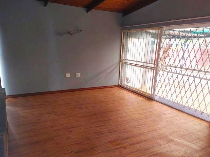 An empty living room featuring laminate flooring, exposed timber ceiling beams, and a sliding glass door with a security gate leading to the exterior.