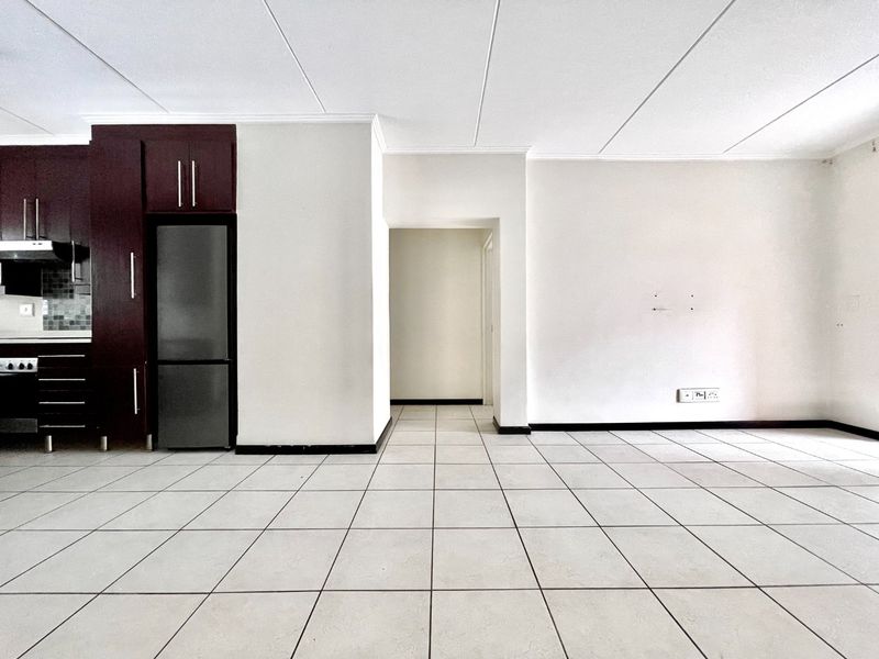 An open-plan living and kitchen area featuring tiled flooring, dark wood-finish cabinetry, and built-in cooking appliances in a clean, vacant space.