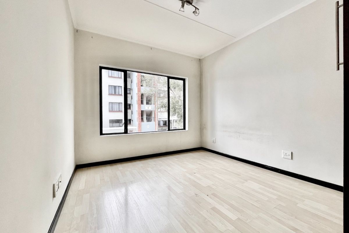 An empty bedroom featuring light-toned laminate flooring, a black-framed window, and neutral walls, providing a blank canvas for personalization.