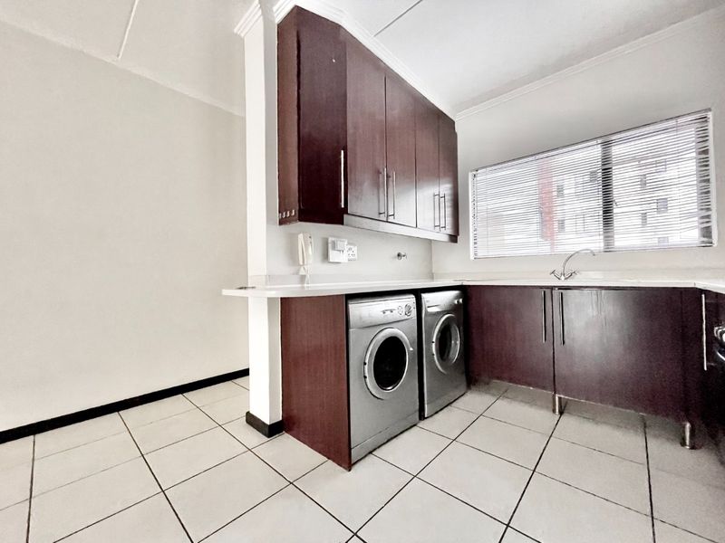 A functional kitchen featuring dark wood cabinetry, laminate countertops, a stainless steel sink, and tiled flooring with space for laundry appliances.
