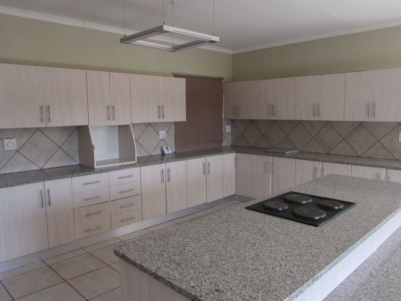 Spacious kitchen featuring light wood-finish cabinetry, granite countertops, a central island with an integrated electric hob, and neutral tiled flooring.