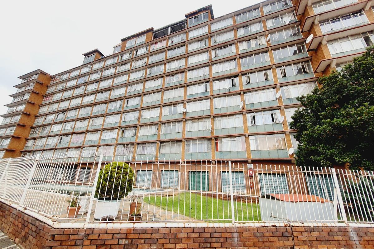 A multi-story apartment building exterior featuring a brick facade, extensive window grid, and secure perimeter fencing with a small ground-level garden area.