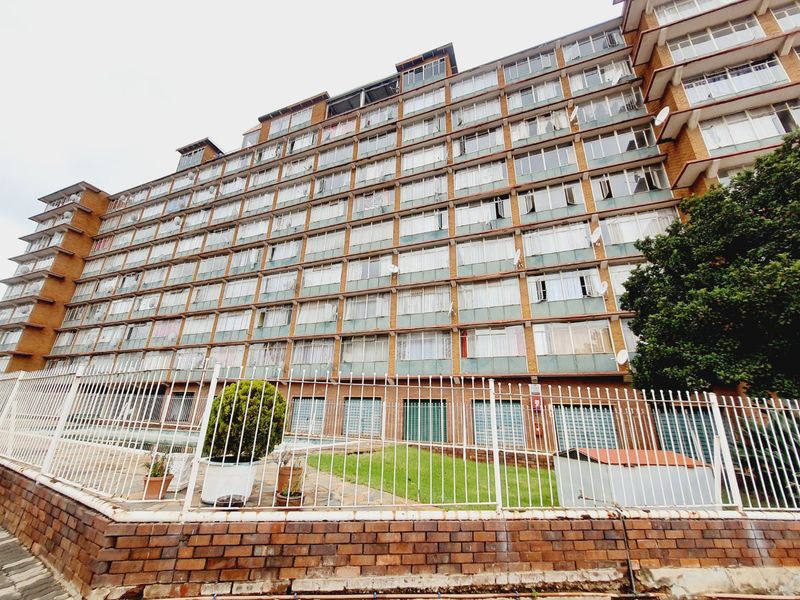 A multi-story apartment building exterior featuring a brick facade, extensive window grid, and secure perimeter fencing with a small ground-level garden area.