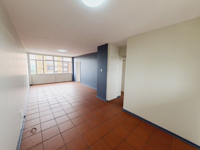 A spacious, empty living area featuring terracotta-style floor tiles, neutral wall finishes, and large windows providing natural light.