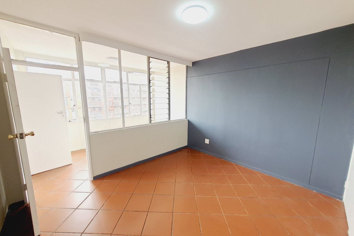 An empty bedroom featuring terracotta-style floor tiles, a dark accent wall, and a glass partition wall with louvre windows providing natural light.
